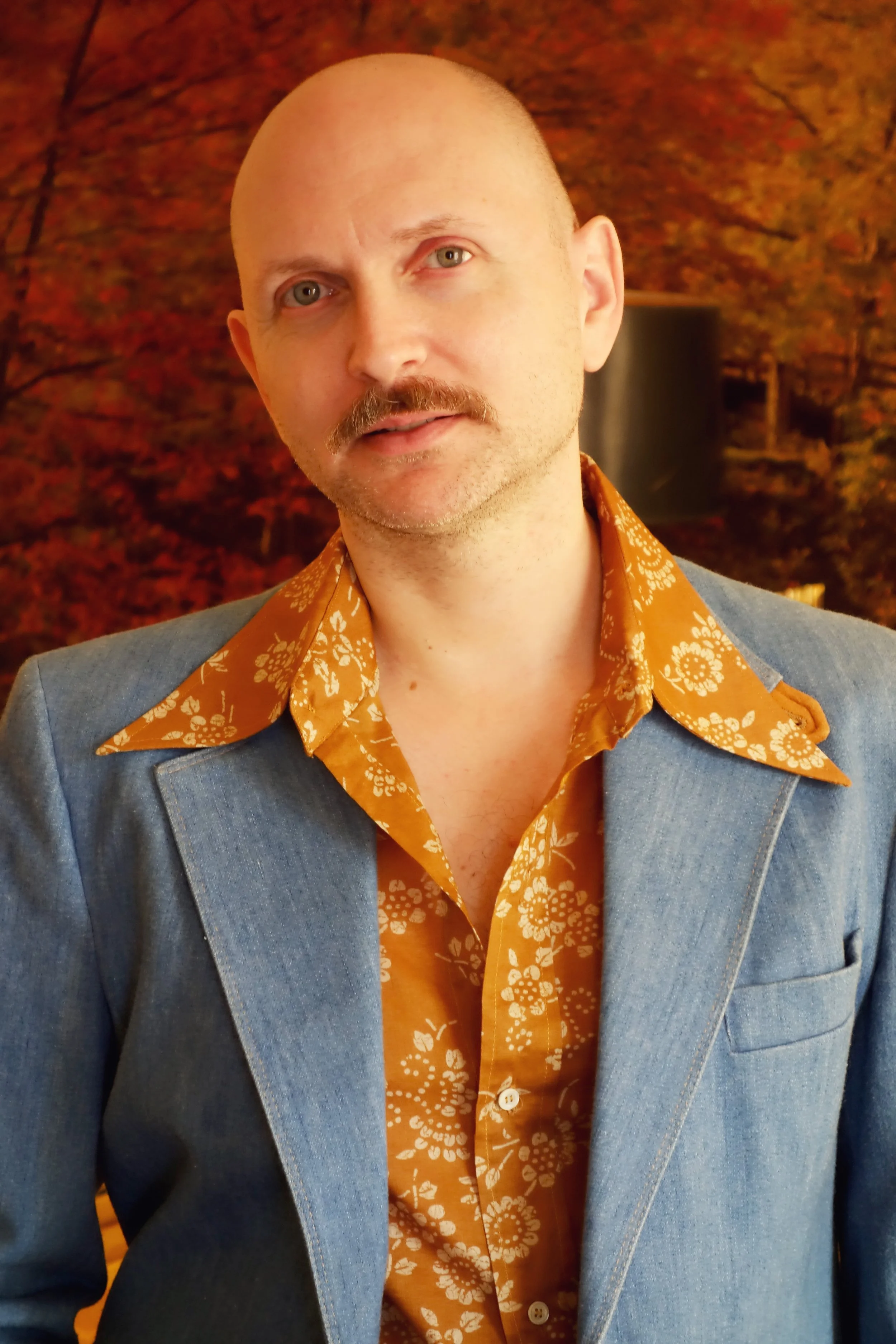Ben Sander with a mustache, wearing a 1970's floral patterned ochre shirt under a denim blazer, stands in front of a background with autumn-colored leaves.