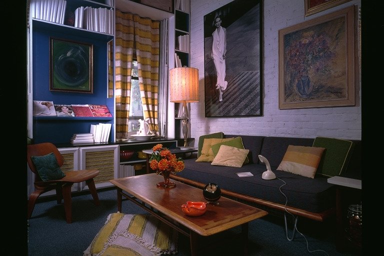 Early 1960's mid-century style living room with a Herman Miller day bed with cushions, a Lane "Acclaim" coffee table with an orange vase of flowers and decorative items, an Eames bent plywood chair with a cushion, a tall lamp, artwork on the walls, a