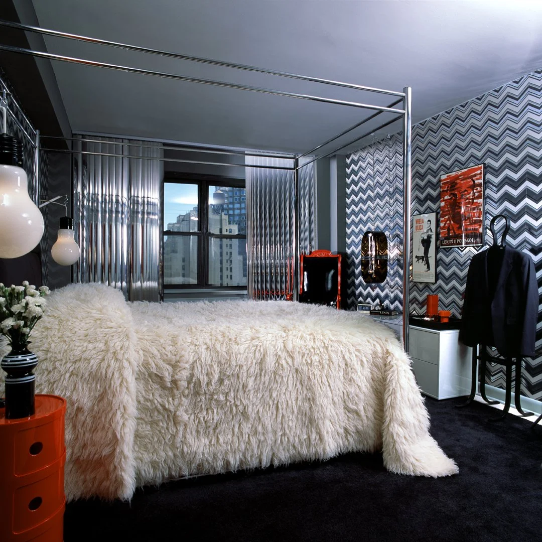 Modern 1970's bedroom with a four-poster bed draped in a fluffy, fur bedspread, located in a city apartment with large windows with silver vertical blinds. Decor includes patterned zigzag wallpaper, black, white and red art posters, and black carpet,