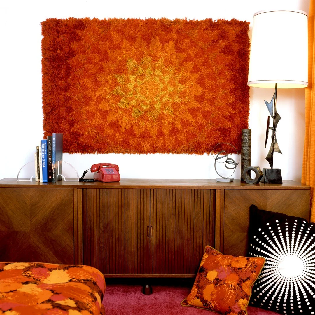 Mod 1970's living room with a large orange shaggy wall hanging, a wooden sideboard, a red push-button phone, books, decorative objects, a floor lamp, a black and white patterned pillow, and an orange-patterned cushion.
