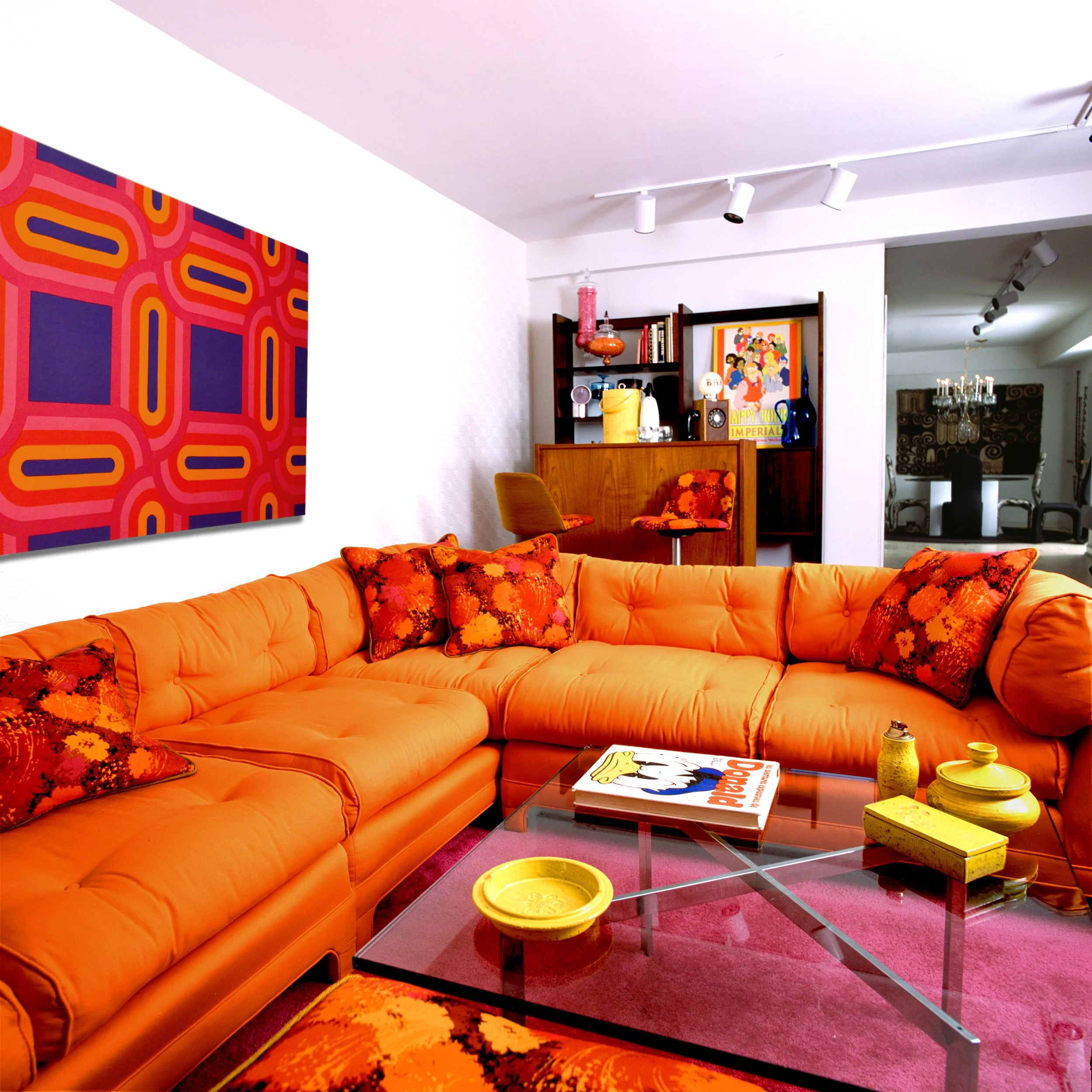 A mod 1970's living room with an orange sectional sofa, colorful throw pillows, a glass coffee table with yellow decorative objects, and a yellow and purple abstract artwork on the wall. There is a bar area in the background with a wooden shelf, book