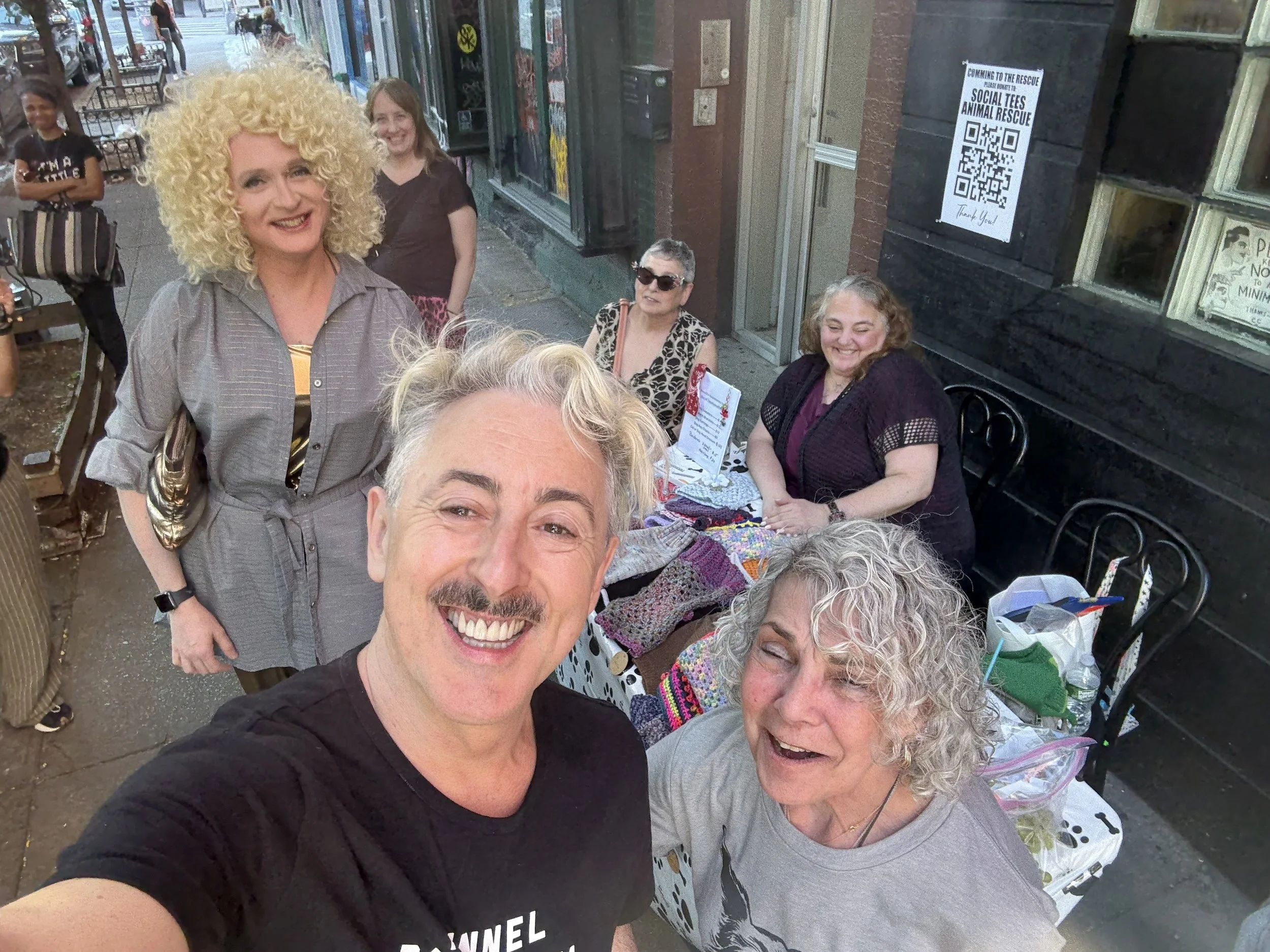 Alan Cumming takes a selfie, with one of the regular attendees of Make It with Brini. Brini Maxwell stands in the background while three other attendees sit at a table nearby, with various objects and bags on the table.