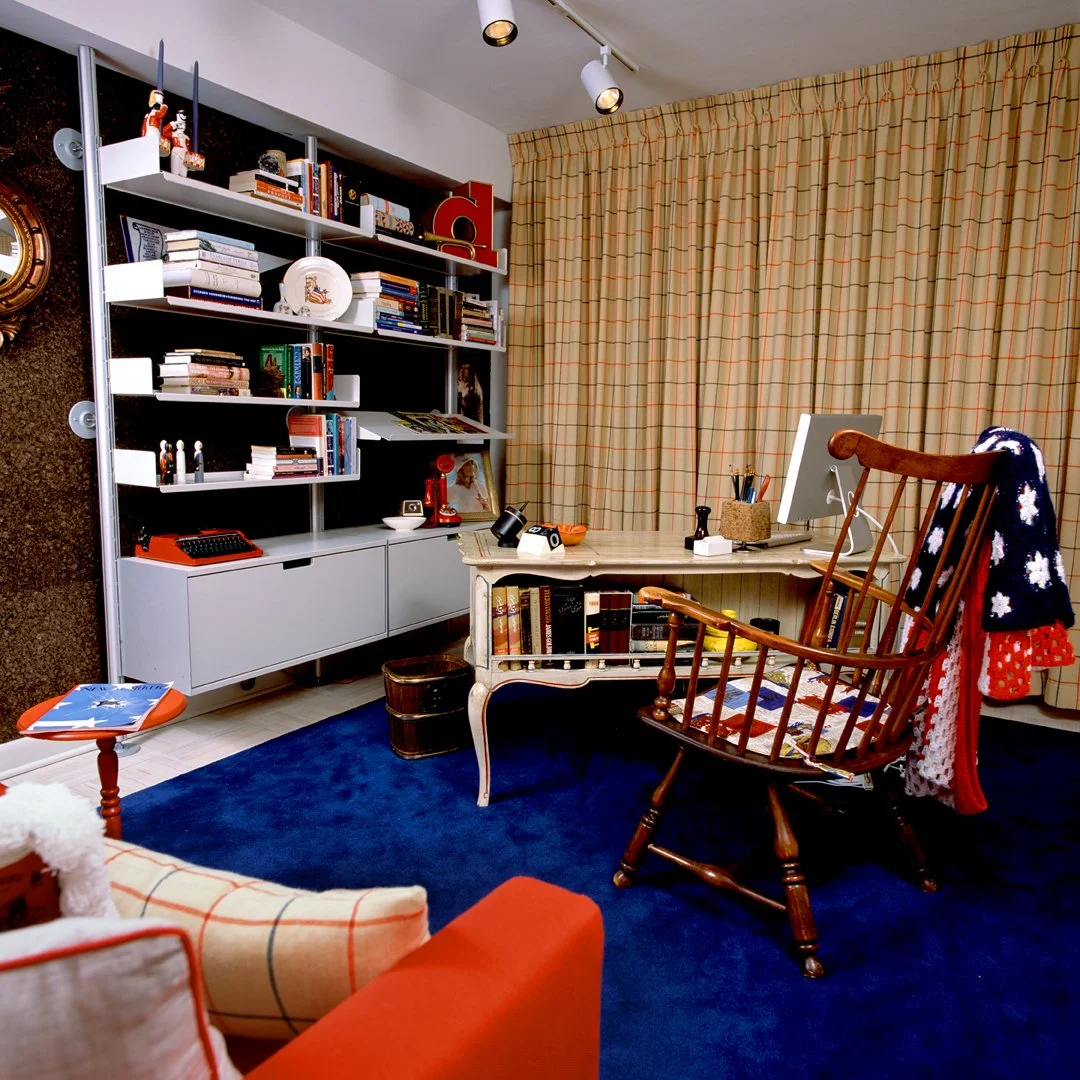 A cozy 1970's mod-meets-colonial home office with a painted and antiqued wooden desk, a Windsor chair, a white modular bookshelf filled with books and decorative items, a computer, and a blue rug. Beige curtains with a red and blue check cover the wi