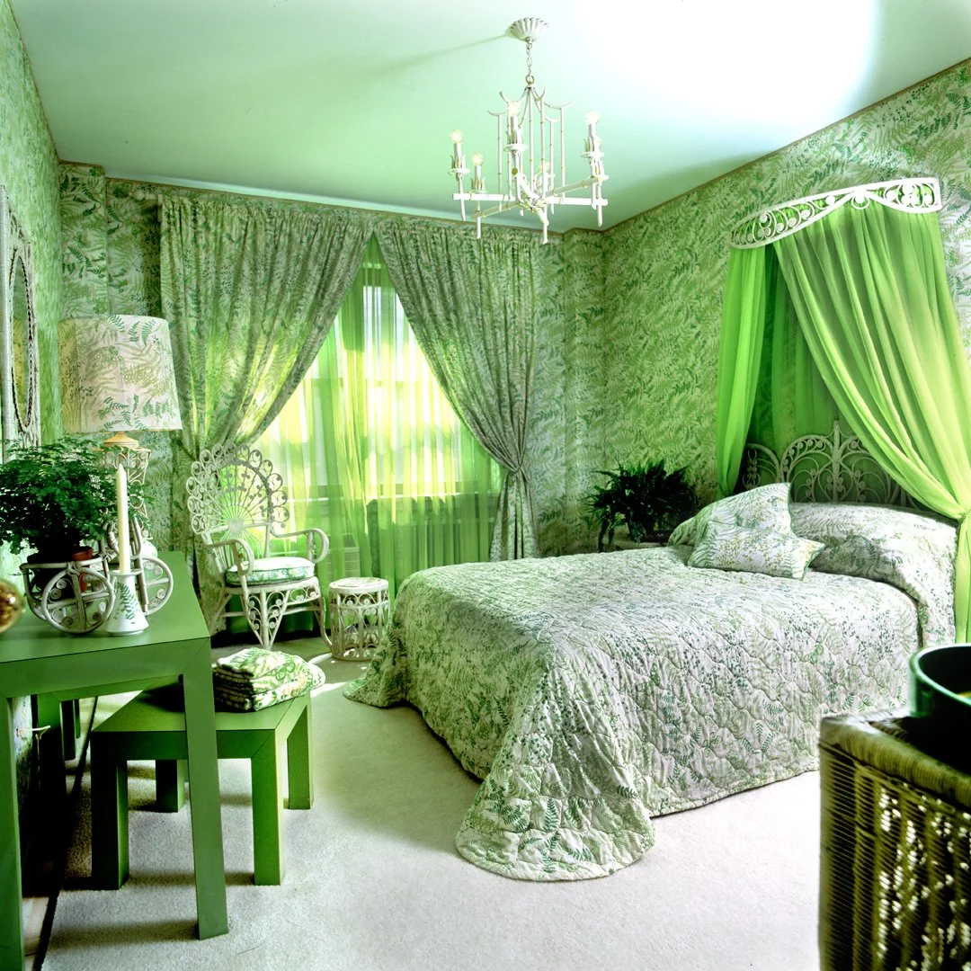 A 1970's jewel-box bedroom decorated in various shades of green, with a canopied bed, curtains, and wallpaper all featuring Vera's green Shadow Fern pattern, wicker furniture and a chandelier hanging from the ceiling.