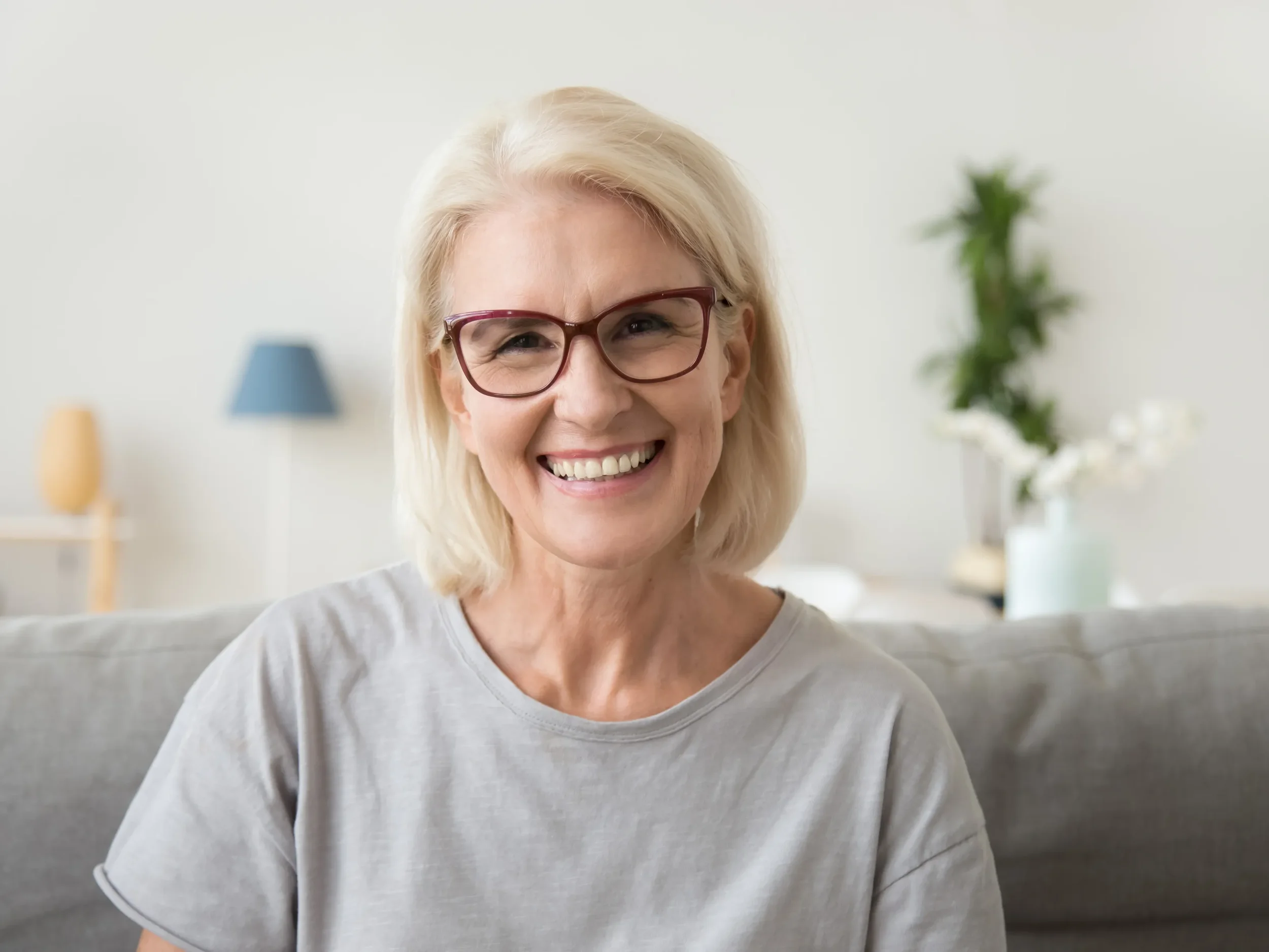 A smiling older woman with blonde hair and glasses sitting on a sofa in a living room.