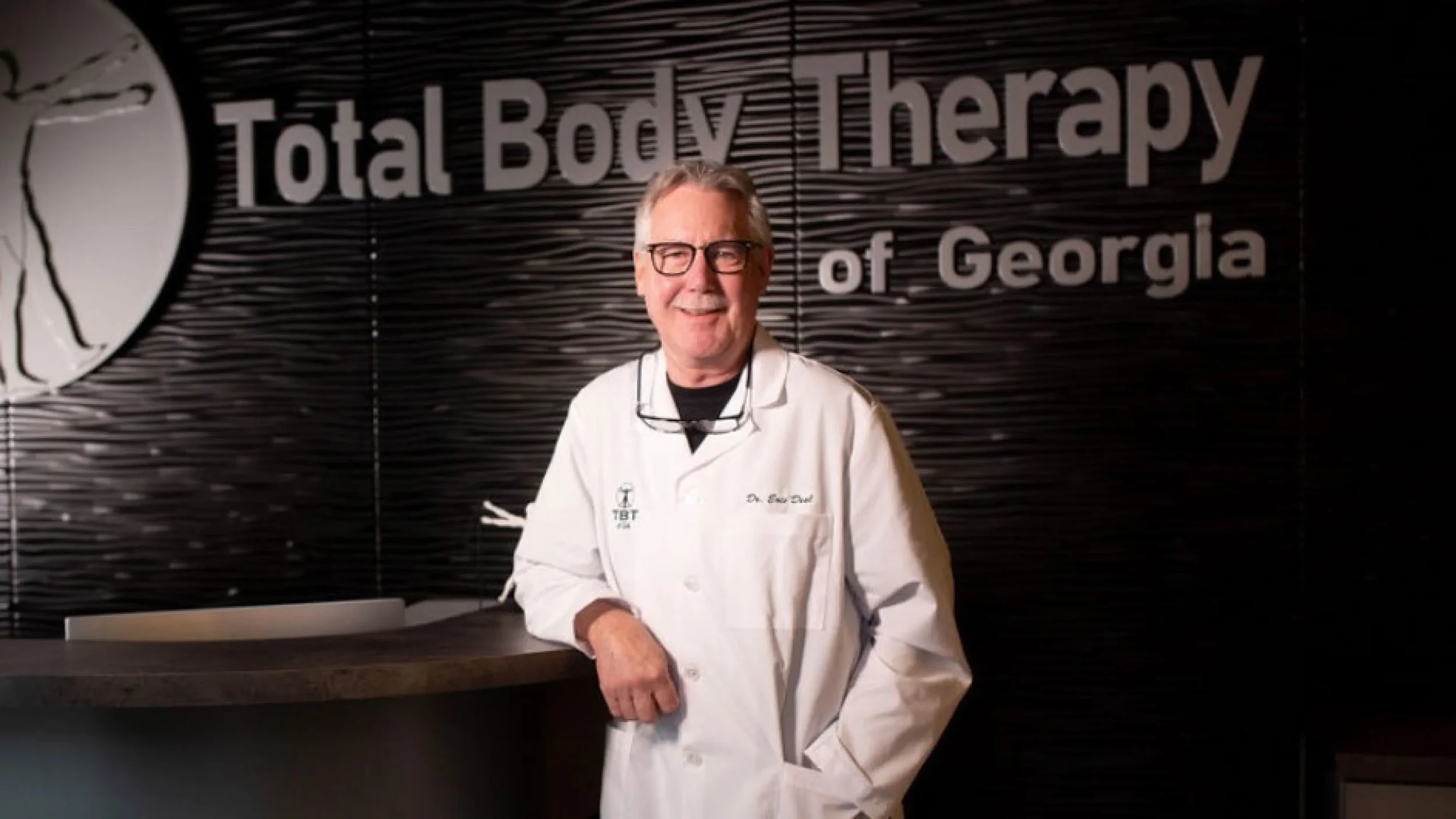 A man in a white medical coat smiling in front of a sign that reads 'Total Body Therapy of Georgia' in a professional lobby or reception area.