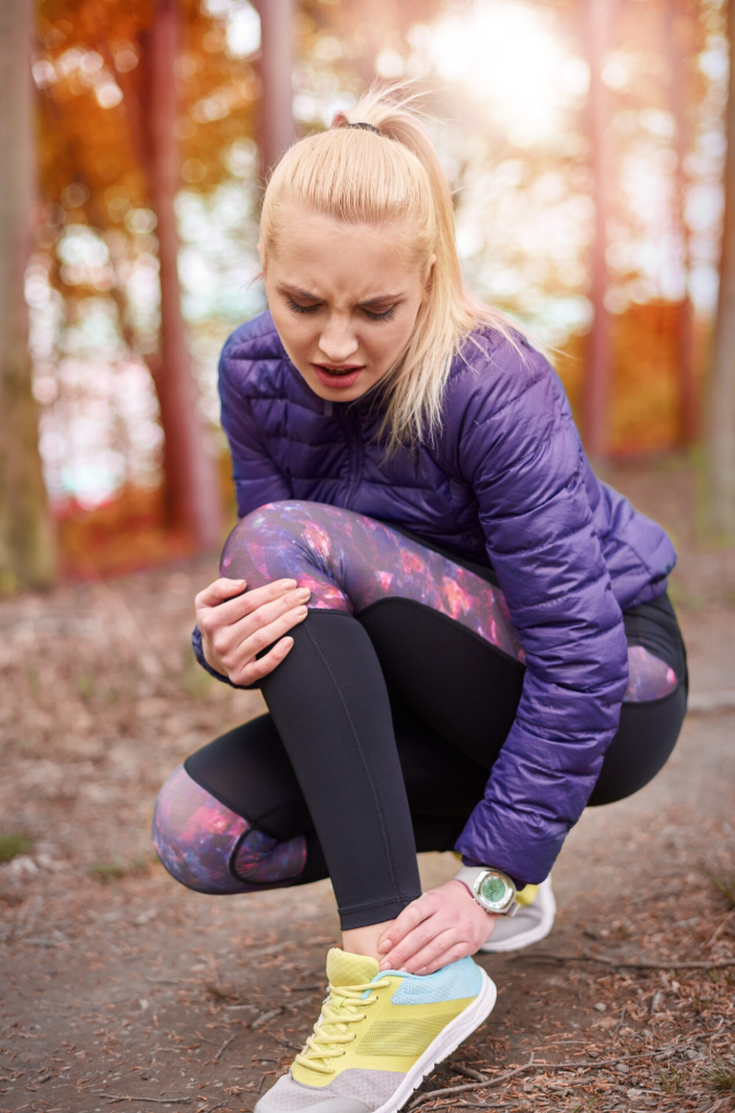 A woman in a purple jacket and galaxy-patterned leggings is crouching in a wooded area, holding her ankle with a pained expression, possibly indicating an injury.