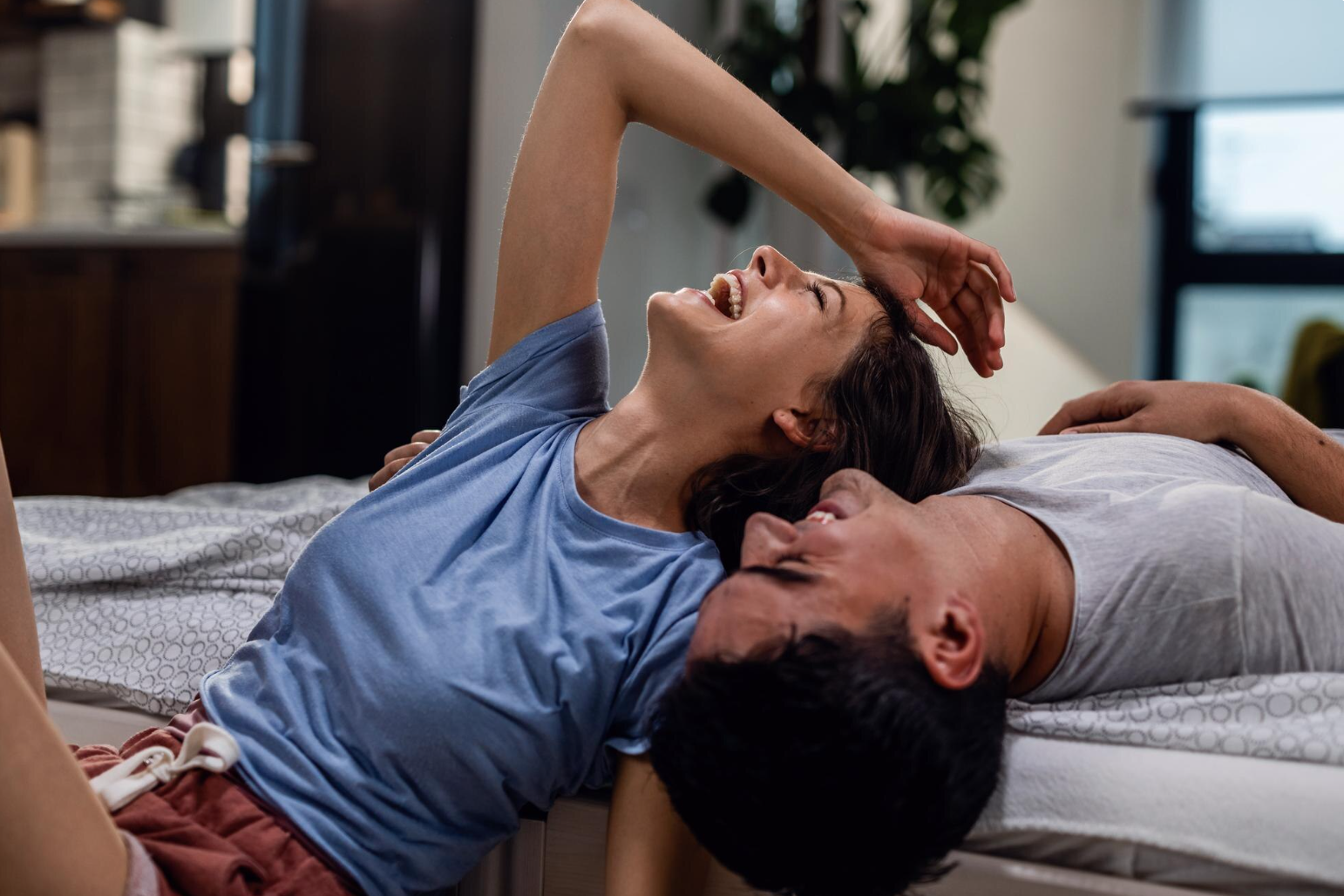 A woman laughing and a man lying on a bed, with the woman resting her hand on her forehead.