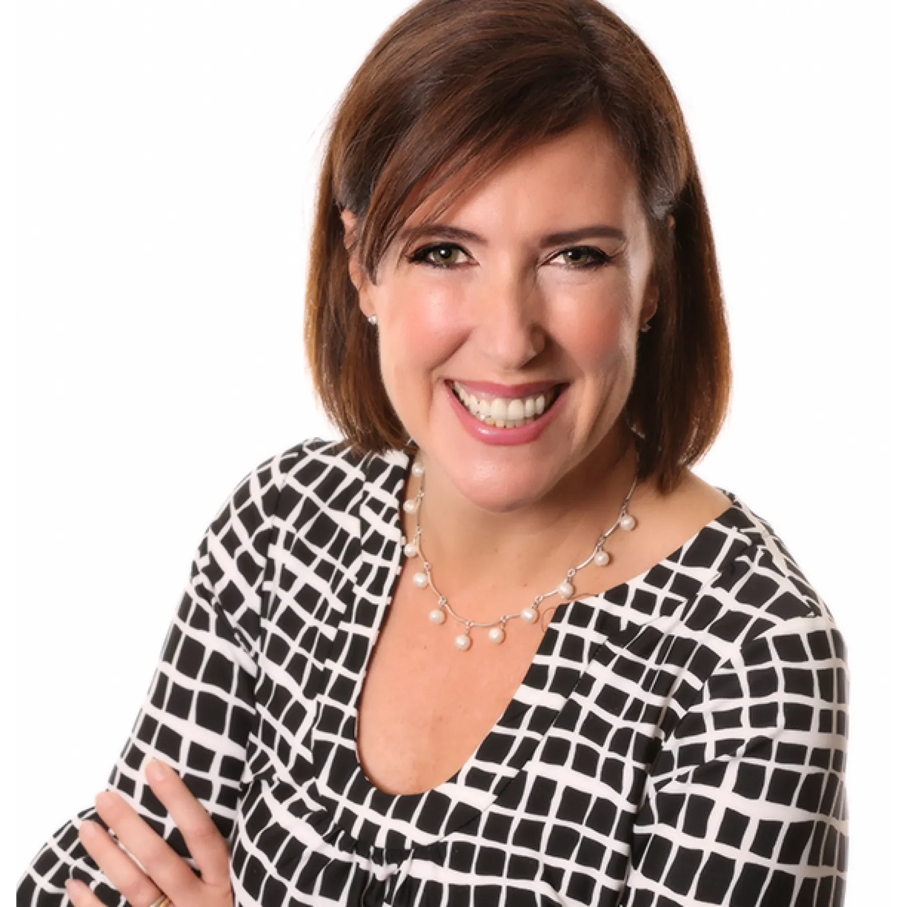 A woman with shoulder-length brown hair, smiling, wearing a black and white patterned top and pearl necklace, with arms crossed against a white background.