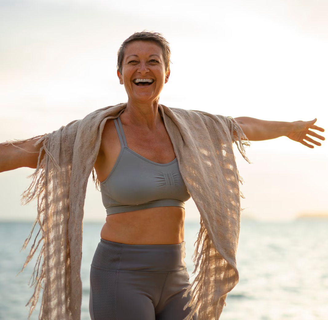 Smiling woman in workout clothes with arms outstretched on the beach at sunset.
