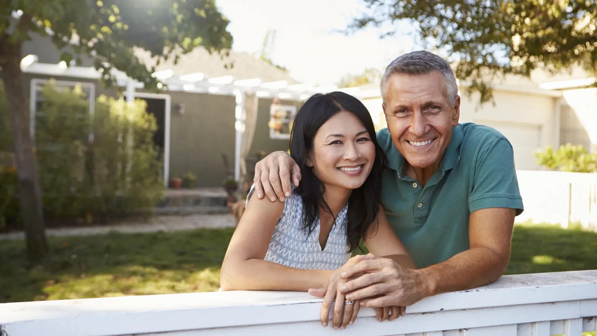 Smiling middle-aged man and woman leaning on a white railing outdoors on a sunny day with trees and houses in the background.