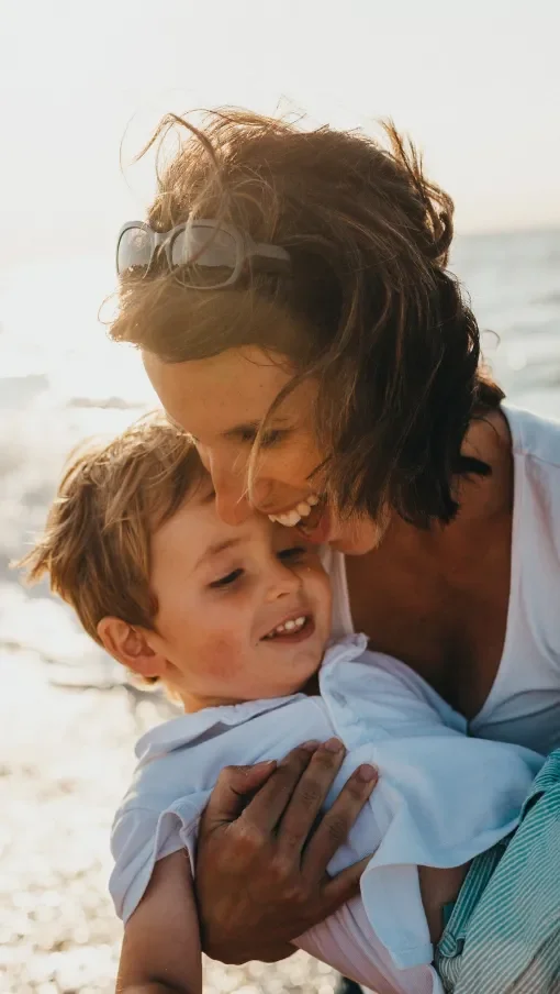 A woman and a young boy sharing a joyful hug on the beach with the ocean in the background.