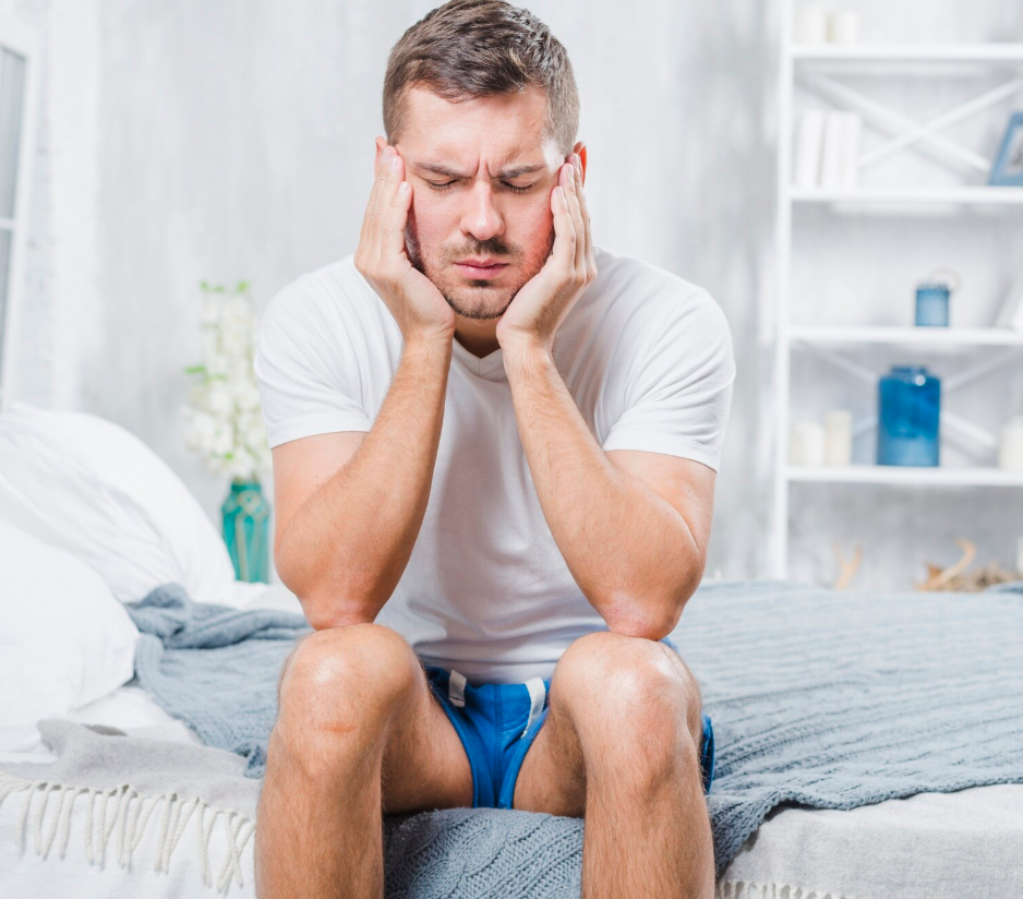 A man sitting on a bed, holding his head in his hands with a distressed expression, appearing to experience a headache or discomfort in a bright bedroom.