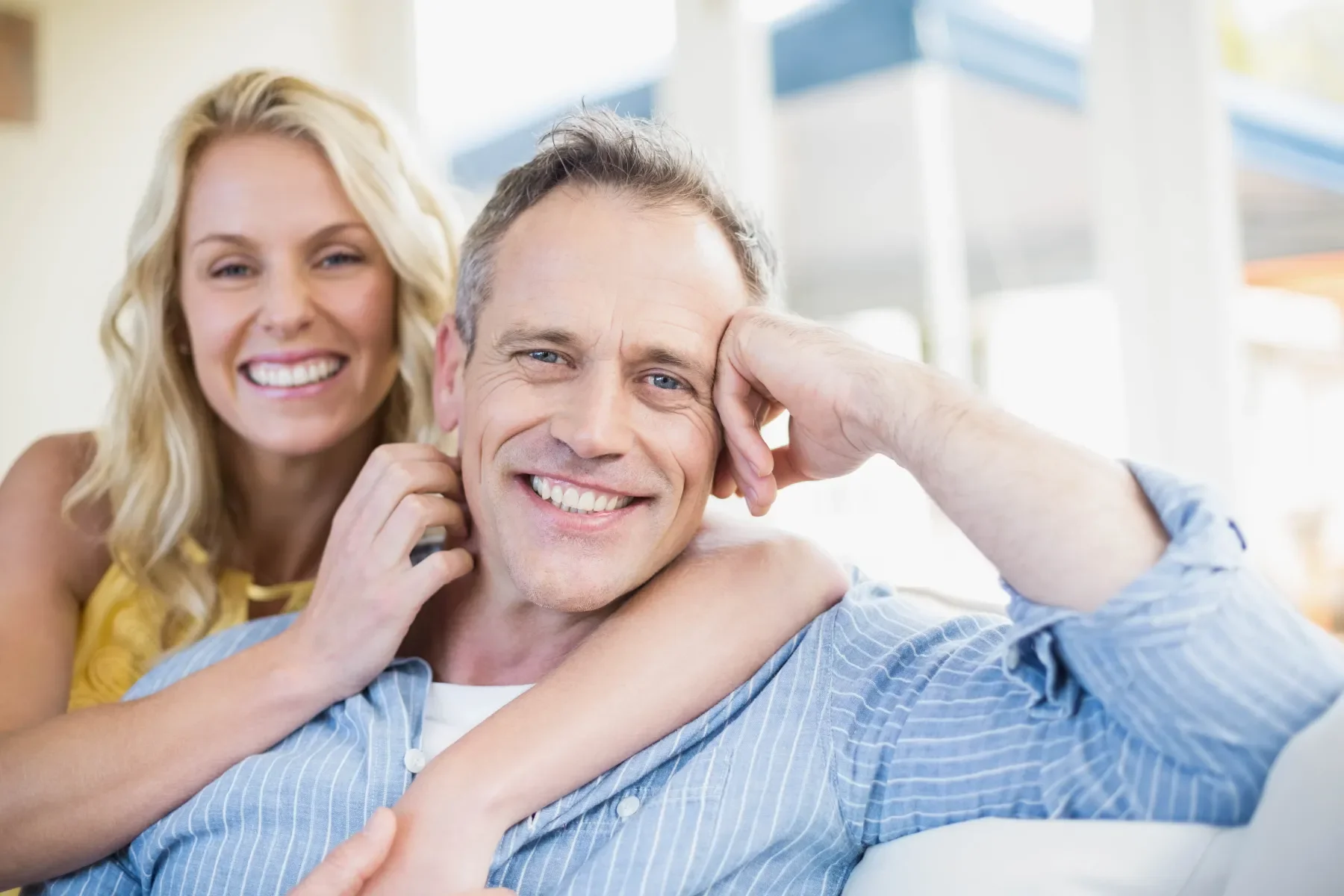 A smiling couple relaxing on a sofa, with the woman behind the man, both looking happy and comfortable.