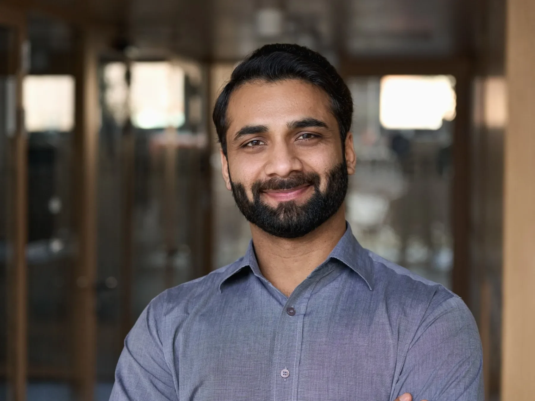 A smiling man with a beard and dark hair, wearing a gray button-up shirt, stands with arms crossed in an indoor setting with warm lighting and blurred background.