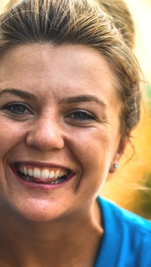 Close-up of a woman with a big smile, showing her teeth, wearing a blue shirt and small earrings, outdoors in natural lighting.