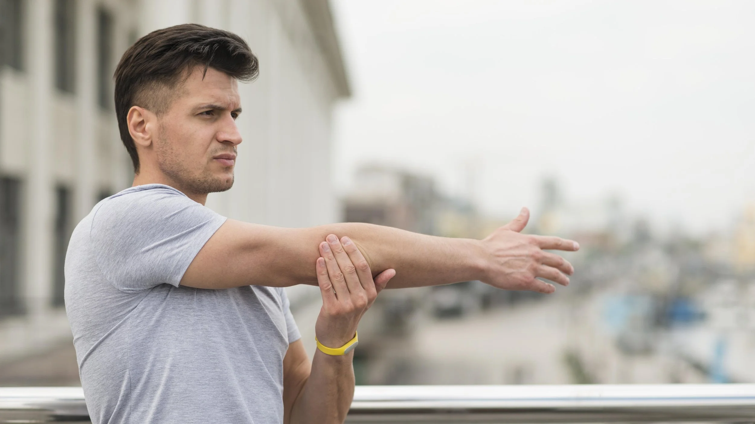 A man stretching outdoors on a cloudy day, wearing a gray T-shirt and a yellow wristband, with buildings and a cloudy sky in the background.