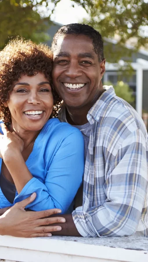 A smiling couple hugging outdoors in a sunny setting with trees and a house in the background.
