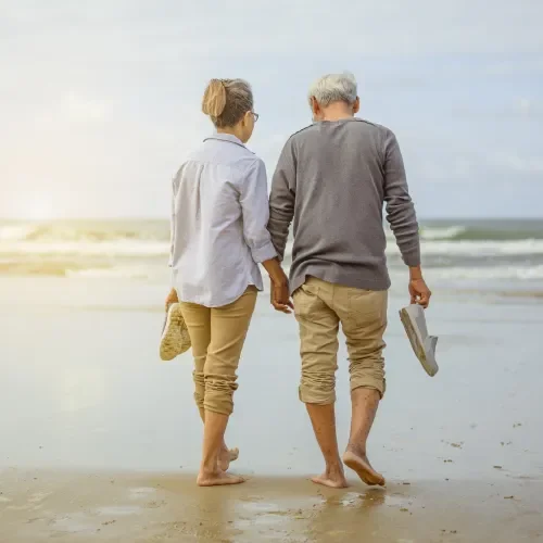 An older couple walking hand in hand on a beach in the evening, with one of them holding a pair of shoes.
