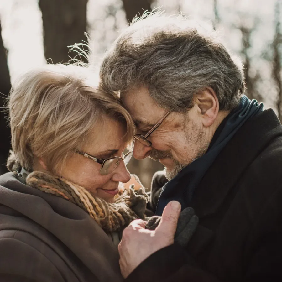 An elderly couple with gray hair and glasses are touching foreheads and smiling, outdoors with trees in the background.