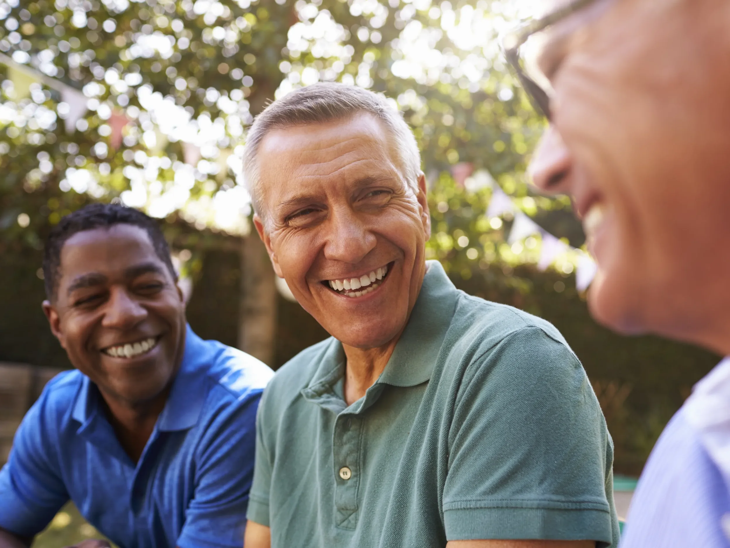 Three men sitting outdoors, smiling and engaging in conversation, with trees and sunlight in the background.