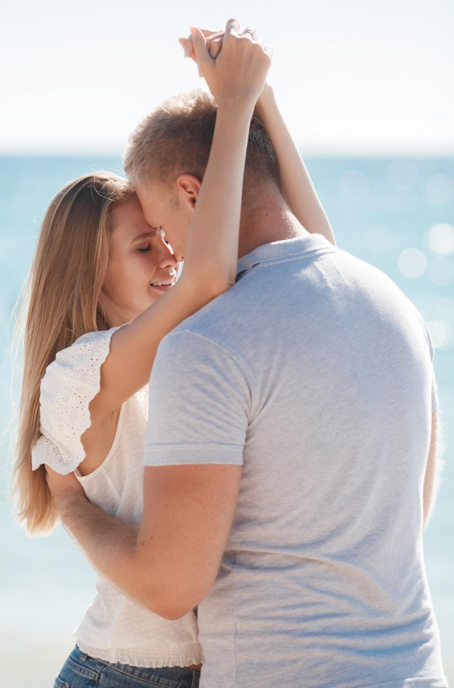 A young couple embraces and kisses by the beach with ocean in the background.