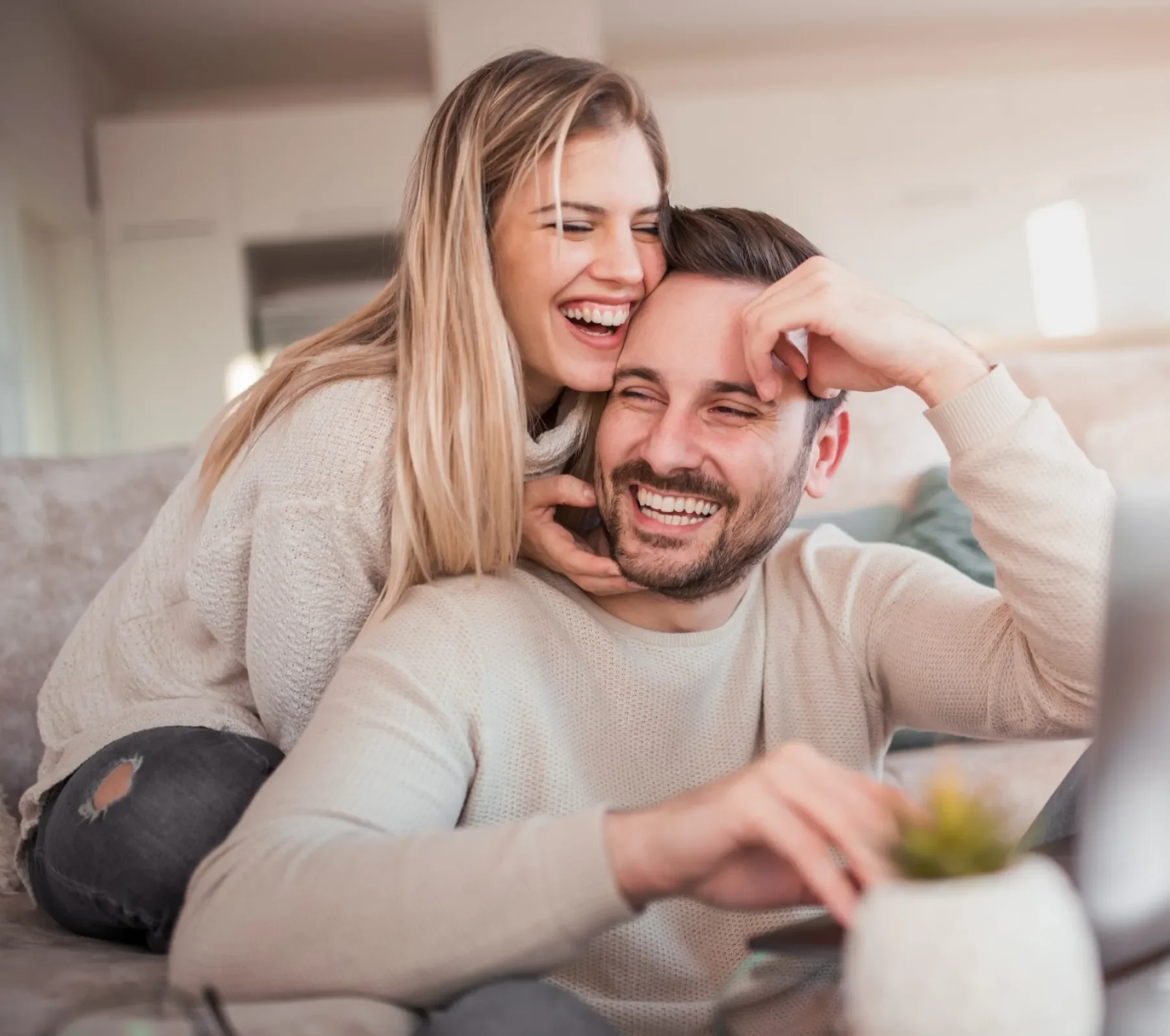 A happy couple laughing and cuddling on a sofa while looking at a laptop.