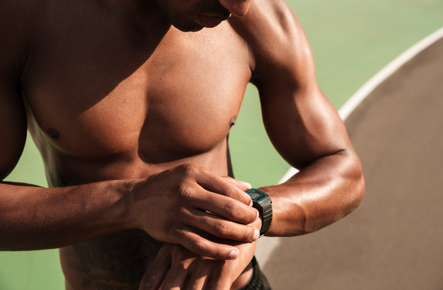 A shirtless man checks his smartwatch with a focus on his muscular arm and upper body, outdoors with blurred green background.