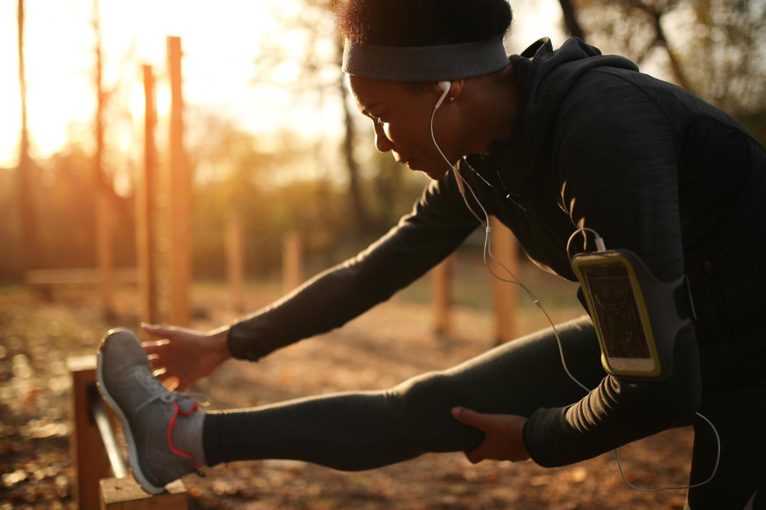 A woman stretching outdoors during sunset, wearing workout clothes, headphones, and a running phone armband.