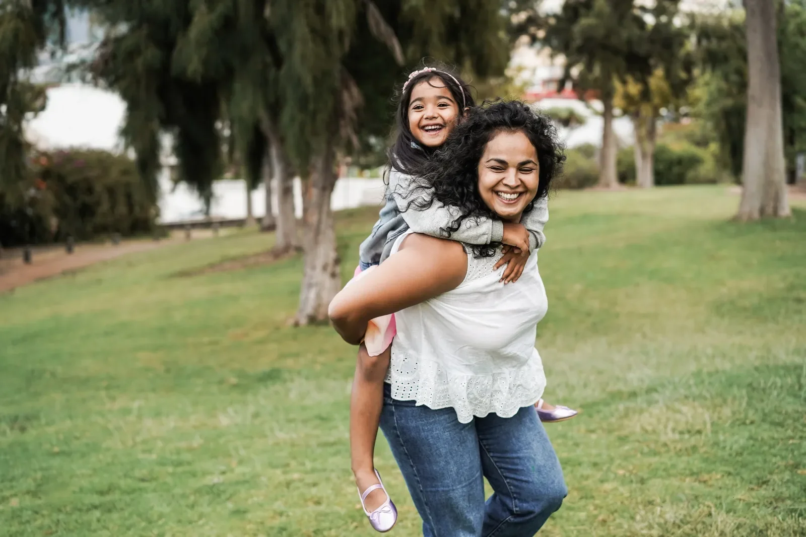A woman giving a piggyback ride to a young girl in a park with green grass and trees in the background, both smiling and enjoying the moment.