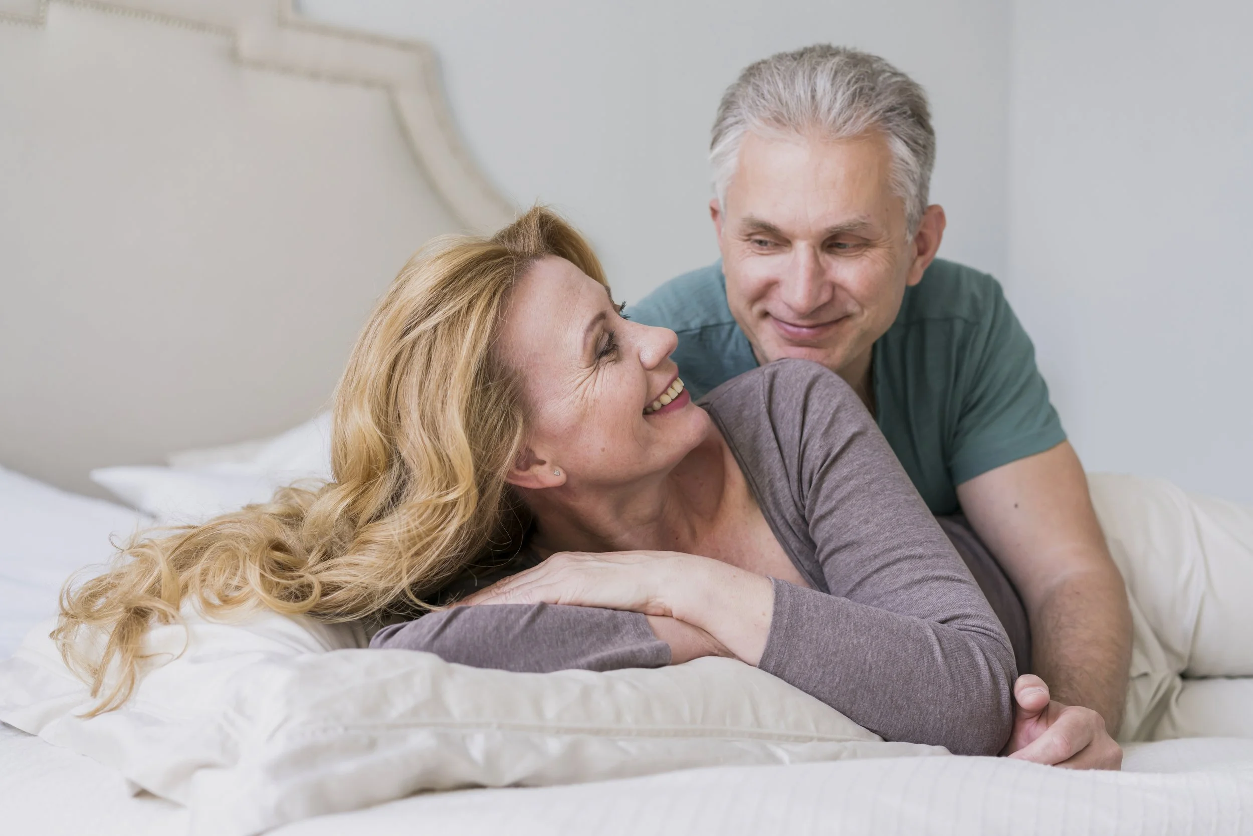 A couple sharing a loving moment on the bed, smiling at each other, with the woman lying down and the man leaning in close.