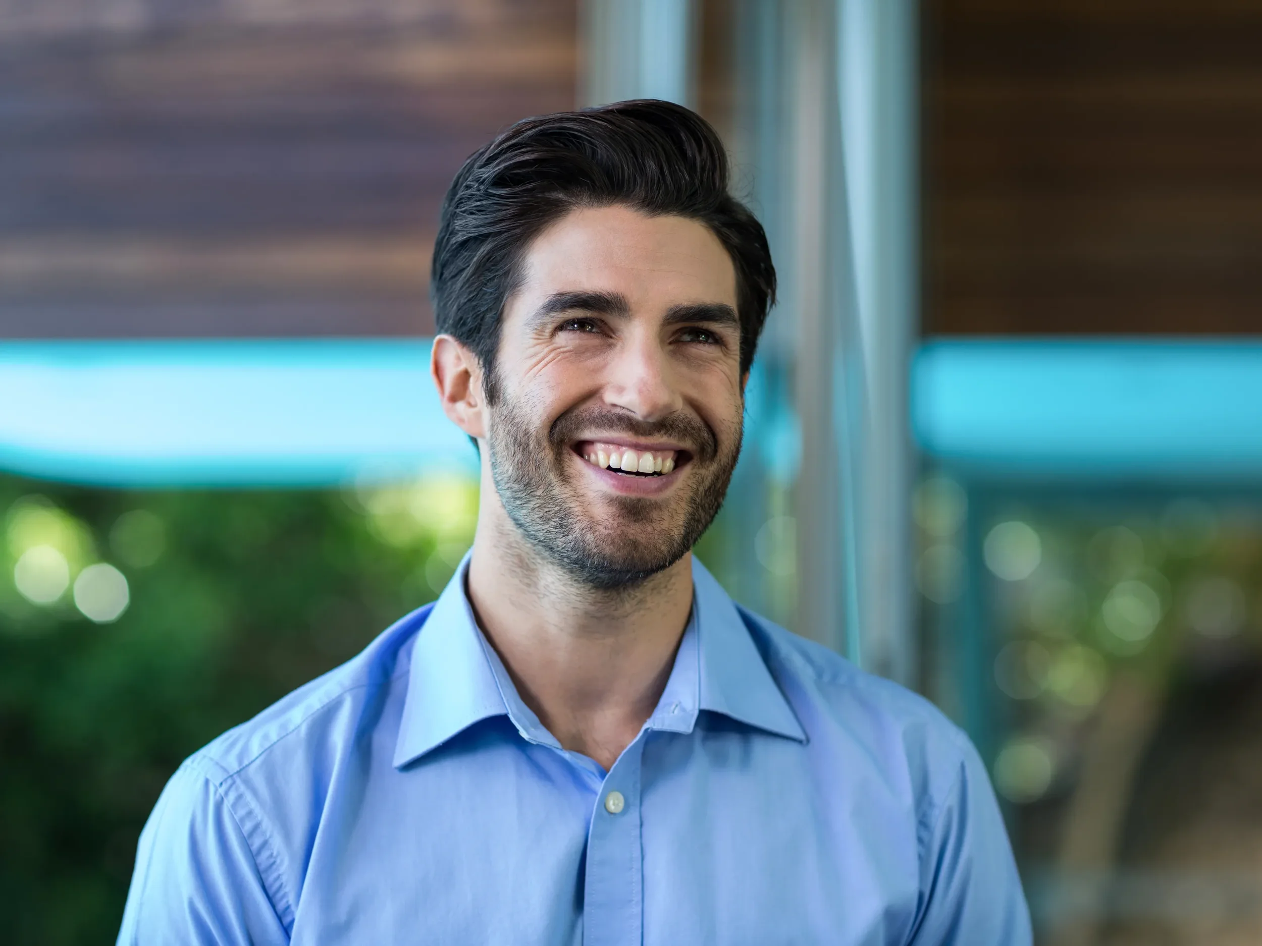 A smiling man with dark hair, a beard, and light skin wearing a light blue dress shirt, standing outdoors with blurred greenery and a structure in the background.