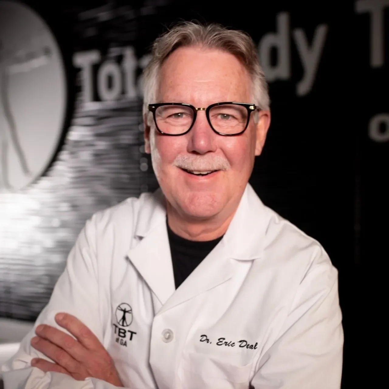 A man wearing glasses and a white lab coat with 'Dr. Erle Deal' embroidered on it, smiling with crossed arms, standing in front of a dark background with a logo and text.