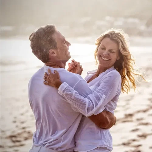 A happy couple dancing and smiling on the beach with the ocean in the background.