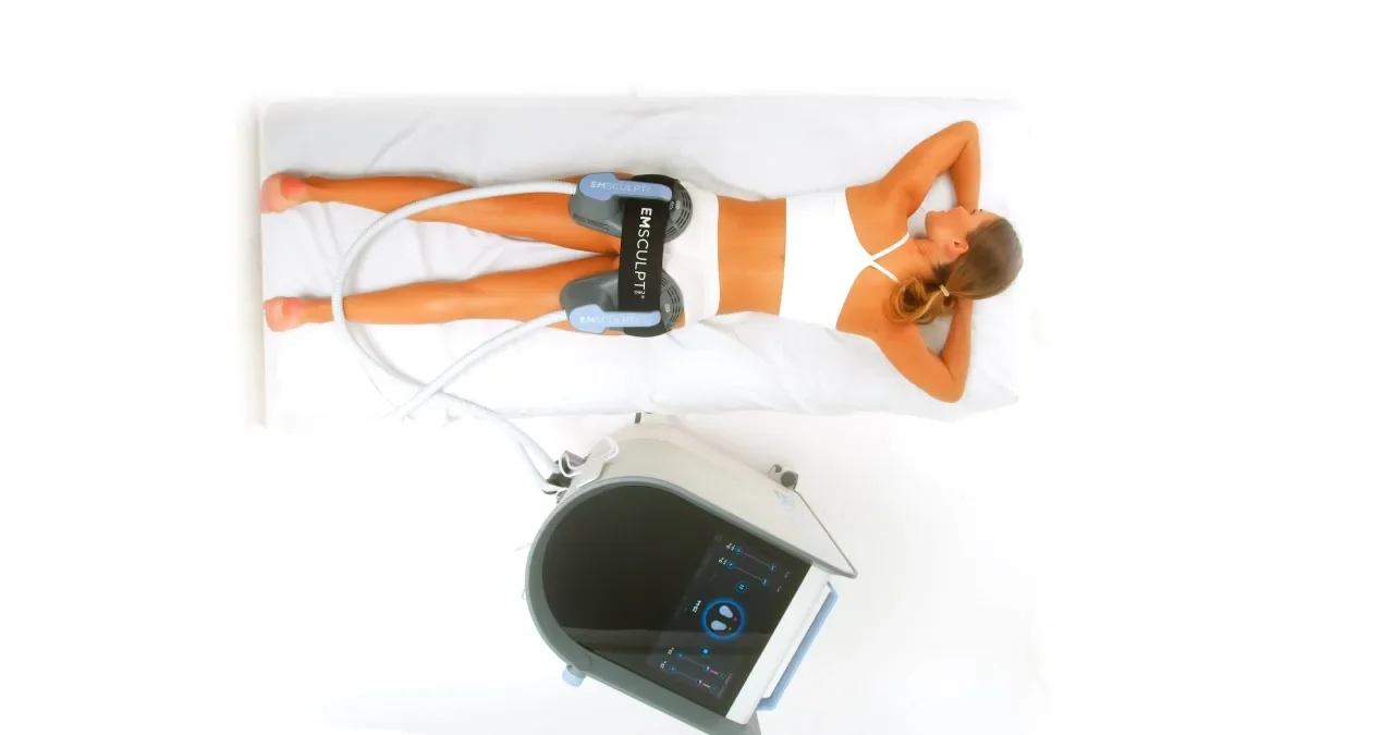 A woman lying on a medical examination table connected to EMSculpt devices for muscle toning, with a medical machine nearby.