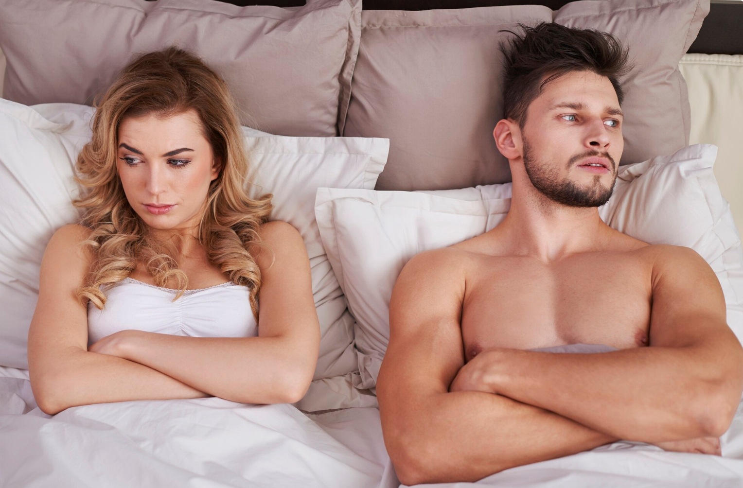 A woman and a man are sitting in bed with arms crossed, looking away from each other.