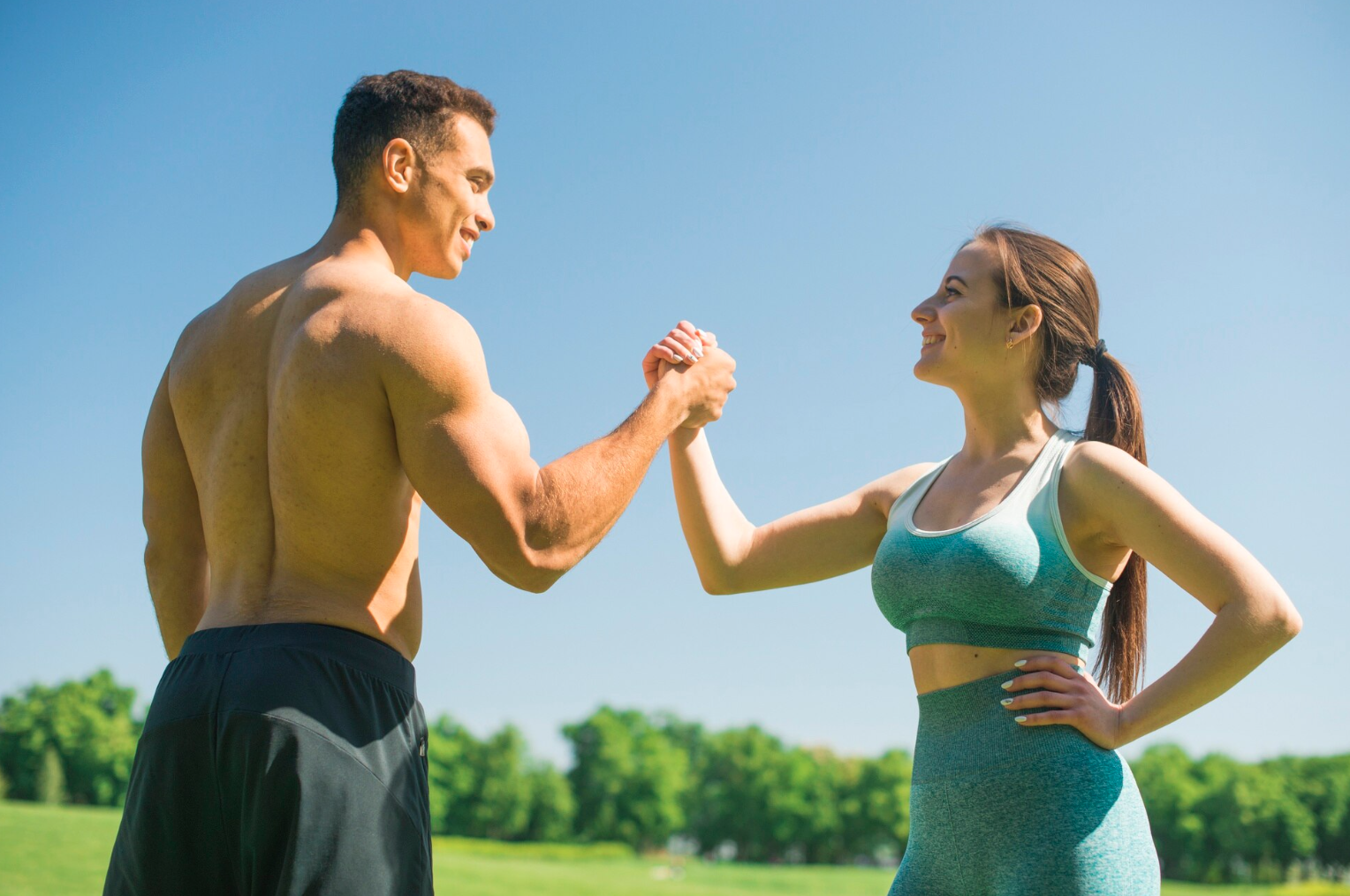 A shirtless man and a woman in workout clothes smiling and arm wrestling outdoors on a sunny day, with green trees and grass in the background.