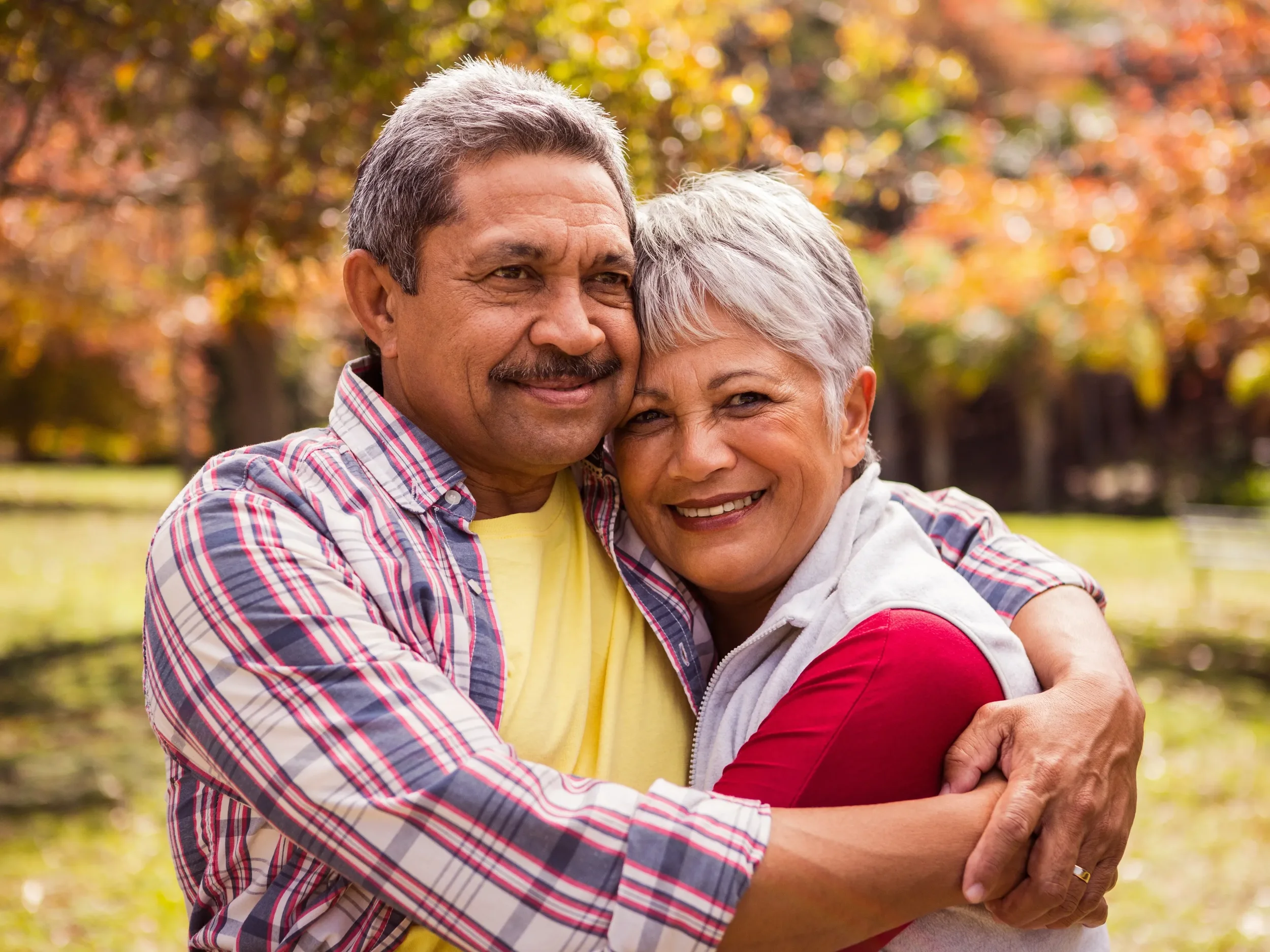 An elderly couple hugging outdoors in a park with autumn trees in the background.
