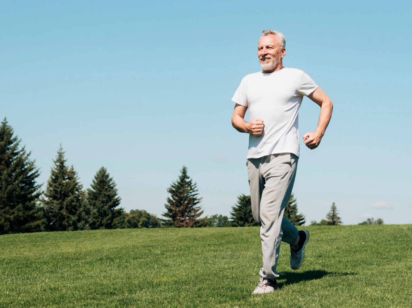 An older man jogging outdoors on a grassy field with trees and a blue sky in the background.