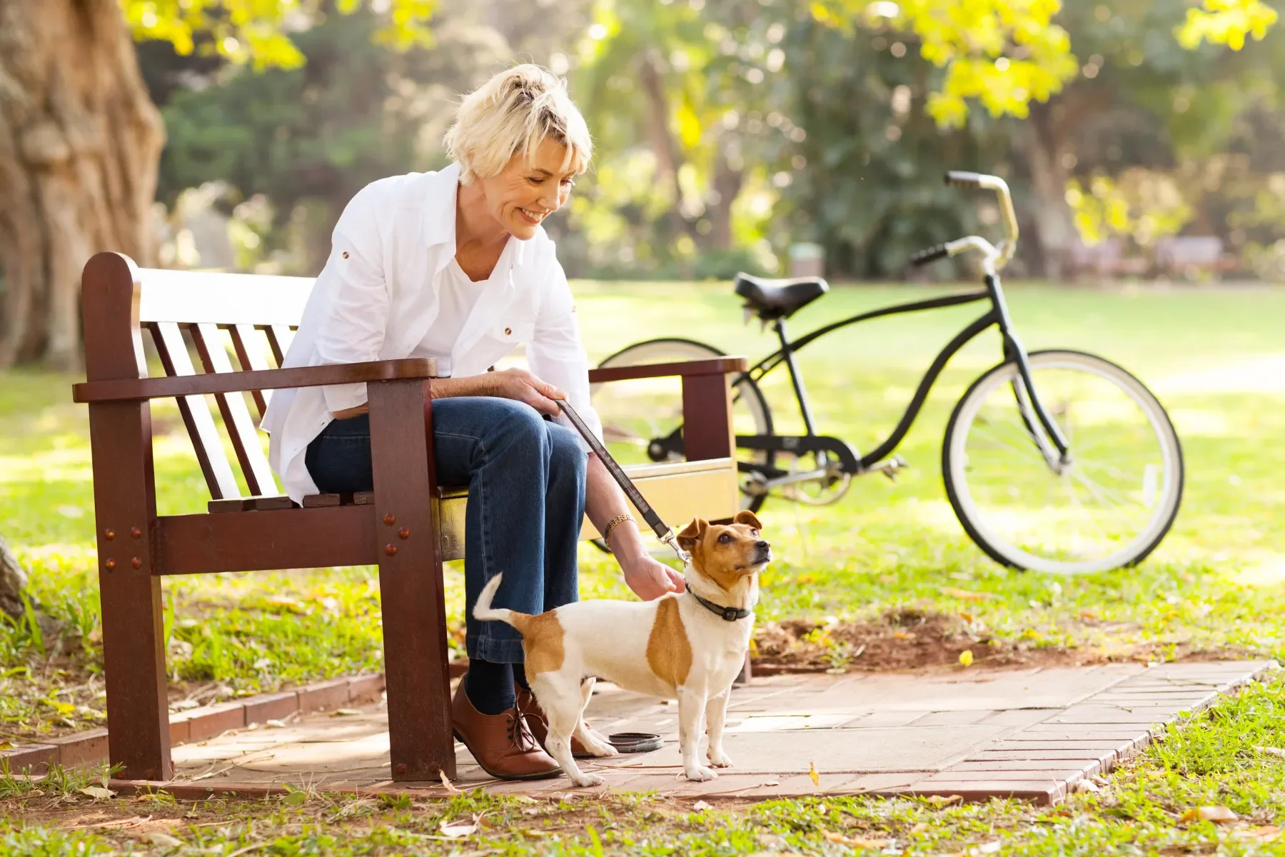 A woman sitting on a park bench with a dog on a leash, enjoying a sunny day with a bicycle in the background.