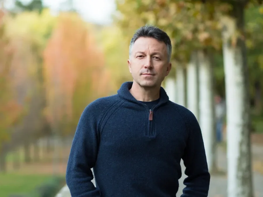 A man wearing a navy blue sweater standing outdoors during fall with blurred trees and pathway in the background.