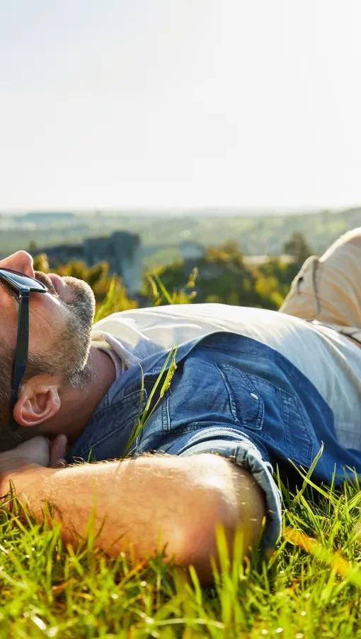 A man lying on the grass in a field, wearing sunglasses and a denim shirt, relaxing in a sunny outdoor setting.