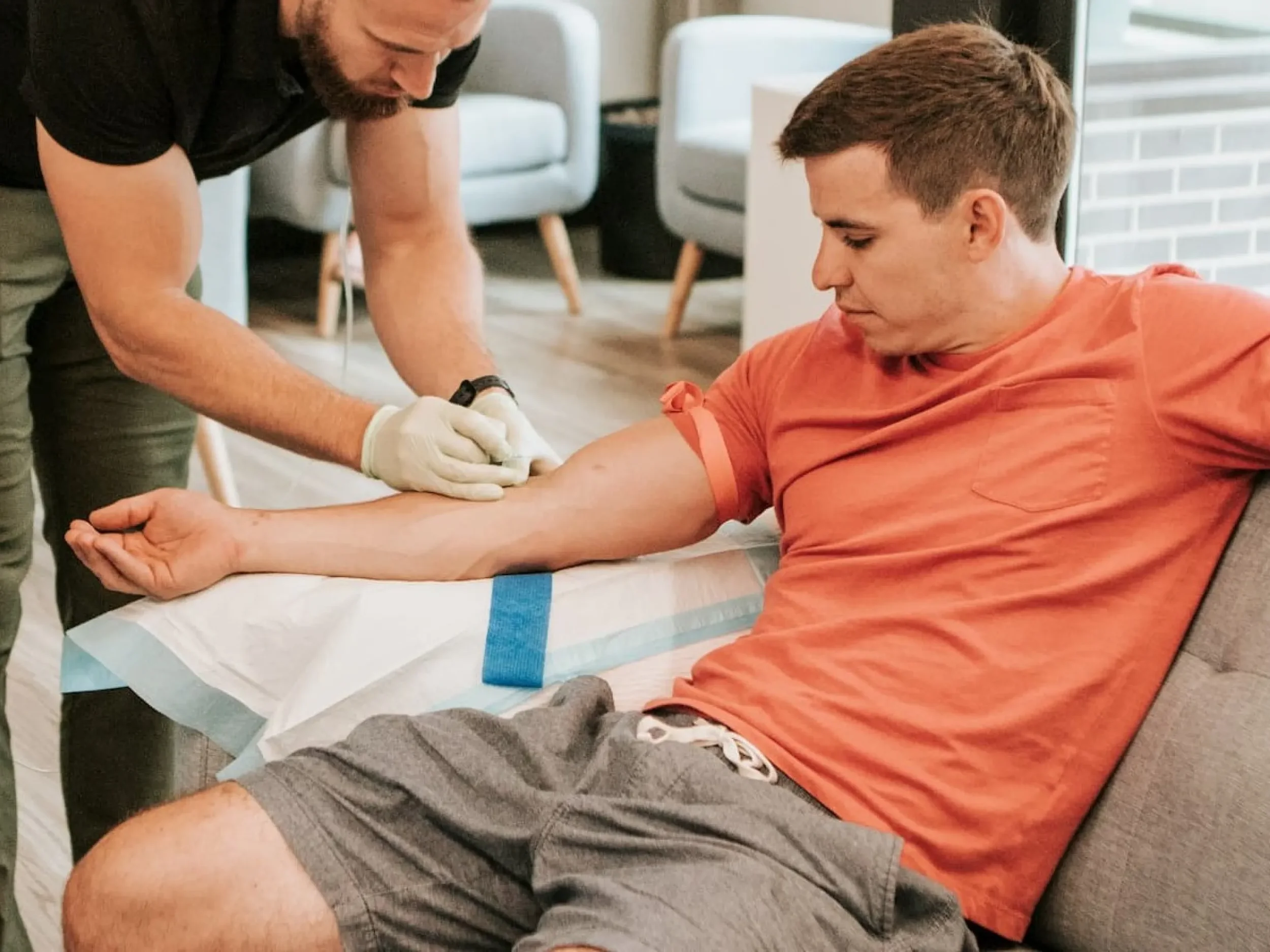 A man is lying on a medical bed, receiving an arm injection from a healthcare professional wearing gloves. The man is wearing an orange shirt and gray shorts, and the healthcare professional is in black attire. The setting appears to be a modern medical facility or clinic.