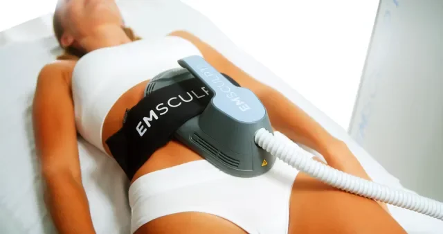 A woman lying on a spa or medical bed receiving treatment with a device labeled 'Emsculpt' placed on her abdomen.