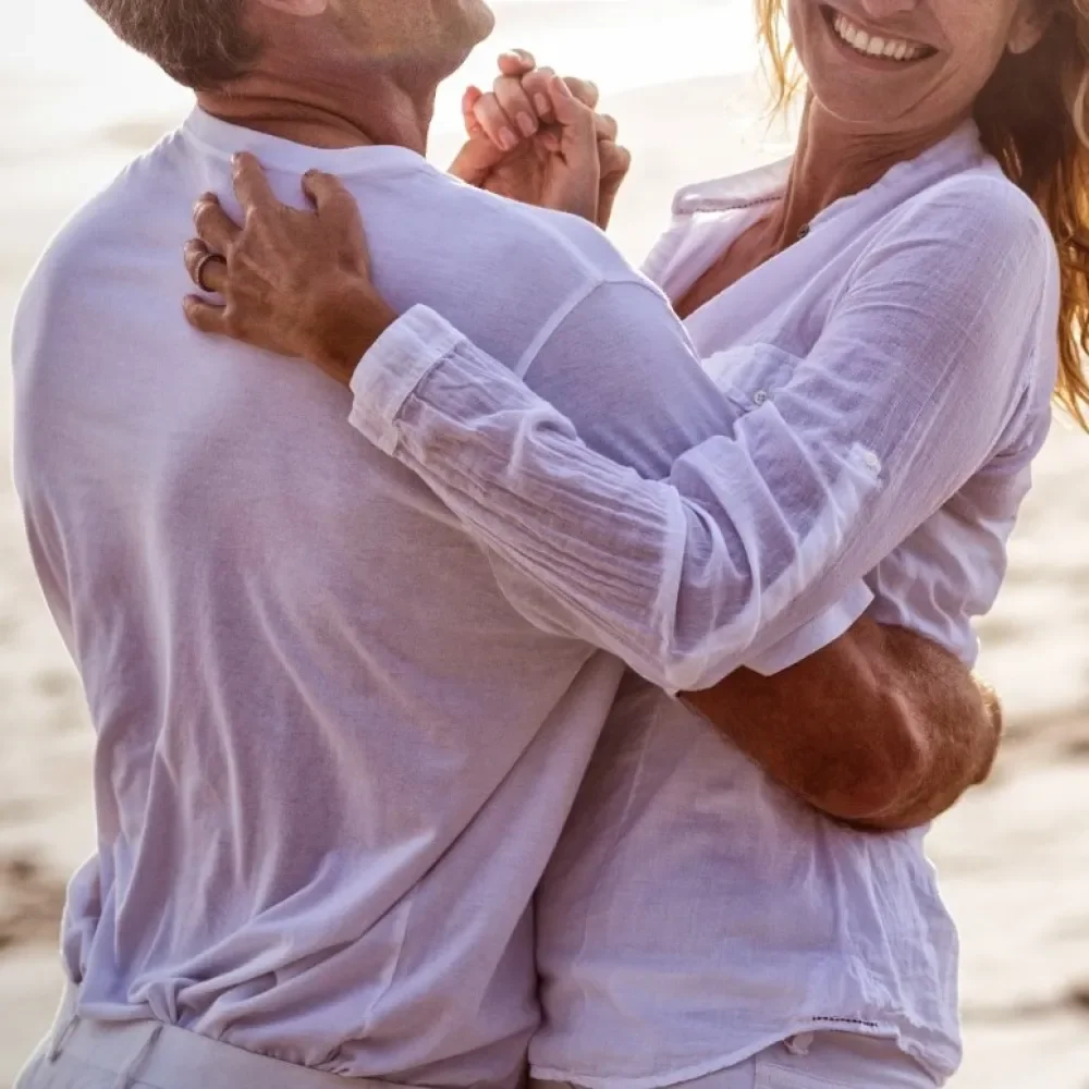 A couple dancing warmly together on a beach, holding hands and smiling, dressed in light-colored clothing.