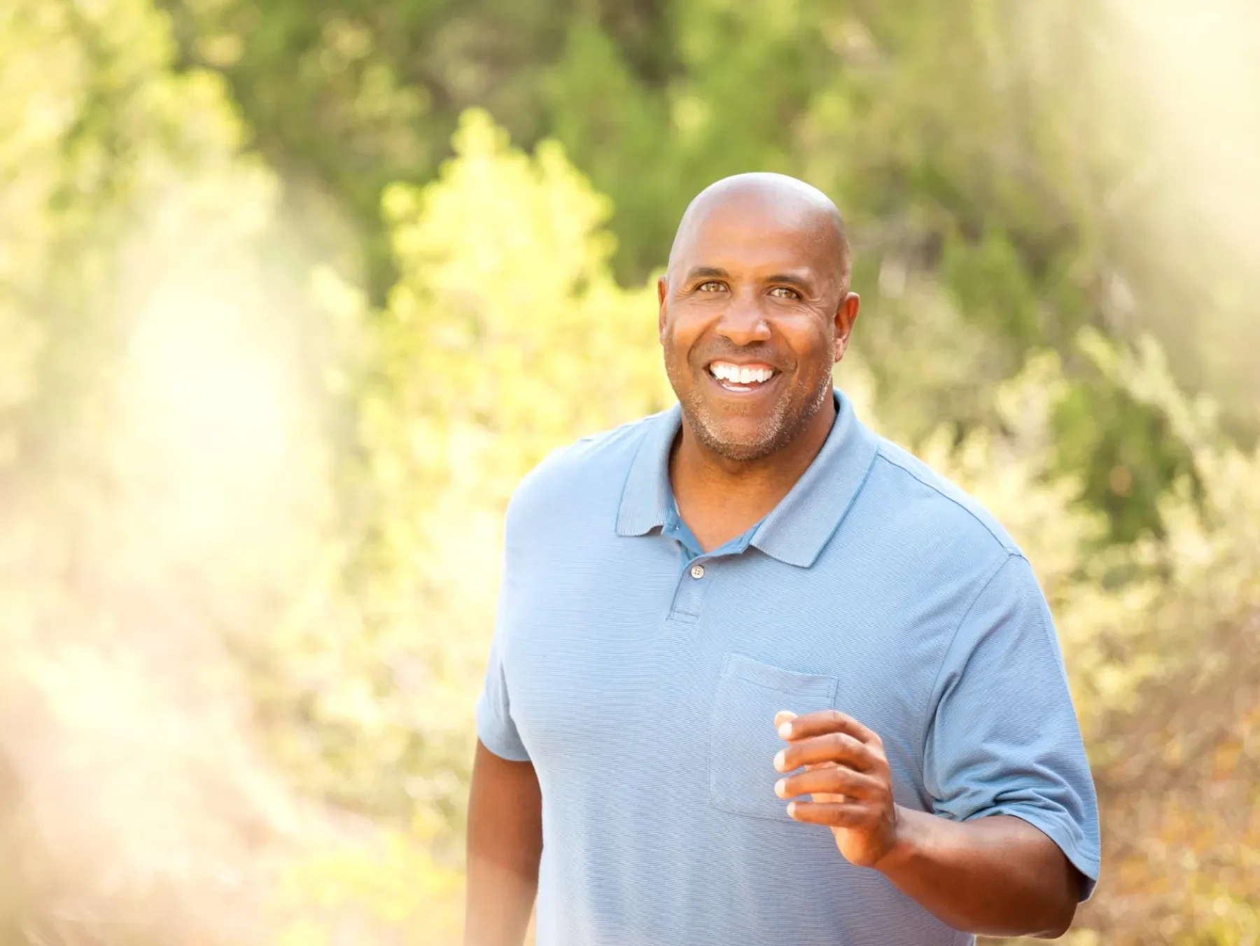 A smiling middle-aged man with a shaved head and a gray beard runs outdoors in a sunny, wooded area.
