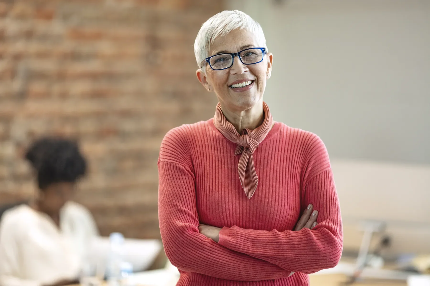 A smiling elderly woman with short gray hair, wearing glasses, a pink ribbed sweater, and a pink scarf tied at the neck, standing with arms crossed in a modern office space.