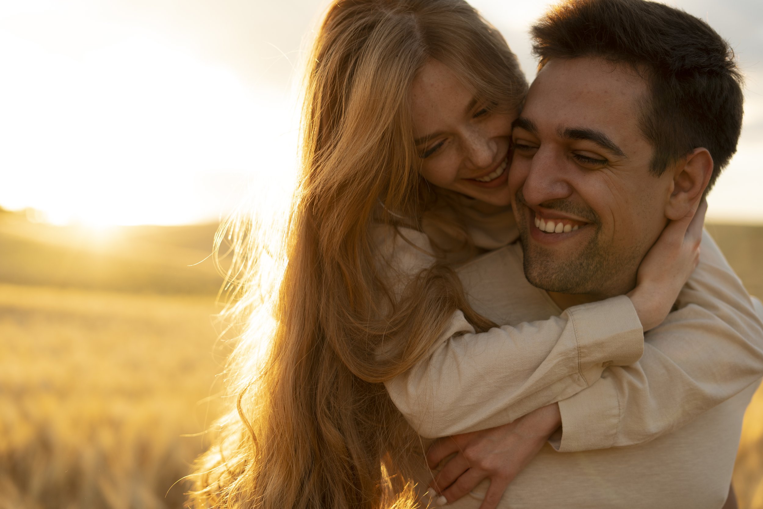 A couple hugging outdoors during sunset, smiling, with golden light illuminating their faces and hair.