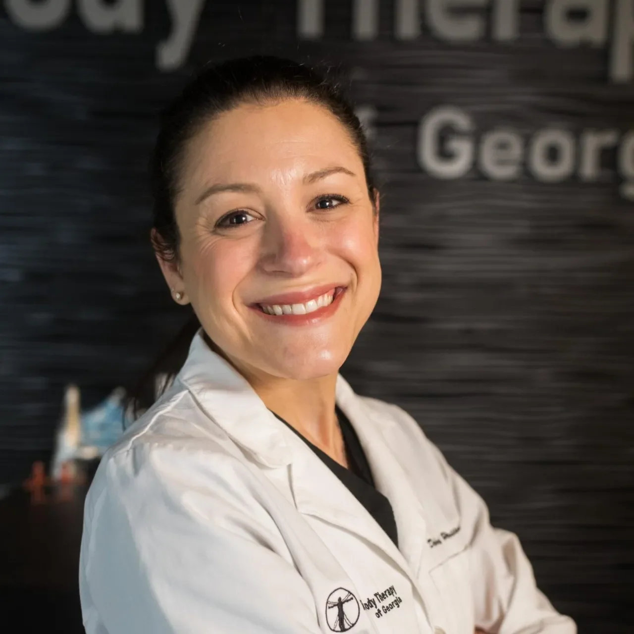 A smiling woman wearing a white lab coat with a logo and text on it, standing in front of a dark textured background with partially visible text.