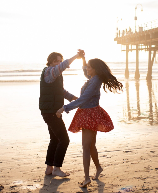 A young couple dancing on the beach at sunset, with the pier in the background.